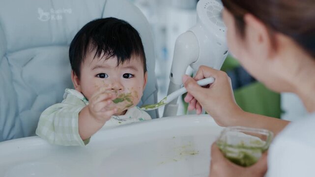 Happy Asian baby boy in high chair smiling at mother while being spoon-fed. Close-up view with natural light in a home setting.	
