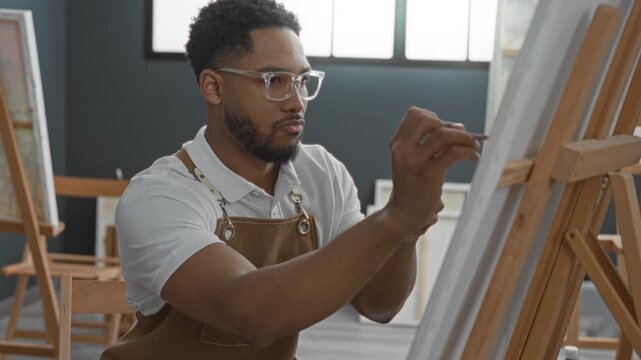Young man painter painting with brush, right hand guiding precise stroke on canvas at wooden easel in studio; creativity concentration.