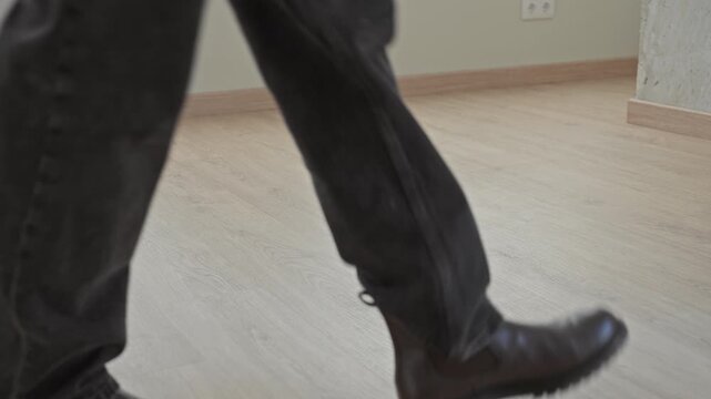 Man walking in leather boots and jeans, stride visible on laminate floor across an empty room inside a building; quiet determination.