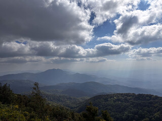 Obraz premium Panoramic view of majestic layered mountains covered in lush tropical forests, stretching into the hazy distance under a dramatic cloudy sky with atmospheric sun rays breaking through.