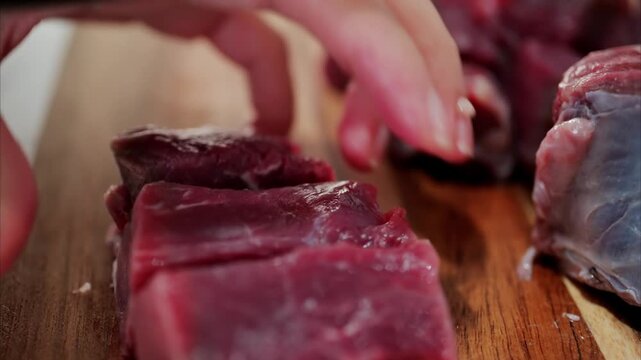 Hands using professional black knife to dice fresh raw beef meat on wooden cutting board in kitchen.