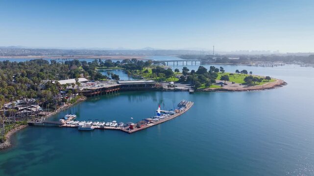 Drone footage of Mission Bay Park island featuring marina with moored boats, lush green parks, palm trees, bridge crossing the bay, and city skyline in the distance on a clear sunny day