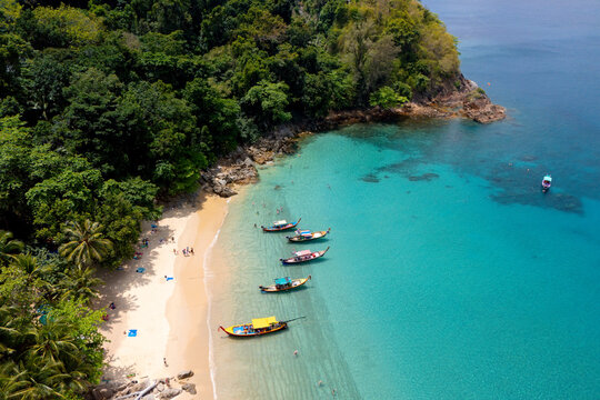 High above Banana Beach Phuket Thailand. See clear turquoise water, multiple longtail boats, and people relaxing on the sand under green palm trees during daytime