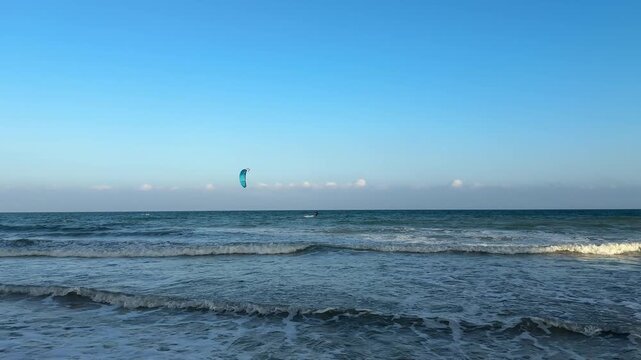 Haikou, Hainan - April 3, 2026: A long-distance 4K shot capturing a kite surfer gliding over gentle waves on a sunny beach, with a red kite against the blue sky in Hainan, China.
