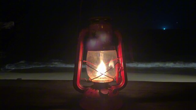 A serene 4K horizontal shot of a classic red kerosene lantern glowing on a wooden dock at night, with rhythmic dark ocean waves and white foam breaking in the background.