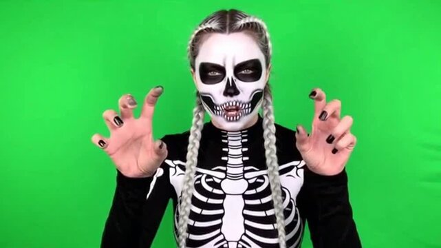 Woman with intricate skeleton face paint and braided white hair wearing a bone-print bodysuit posing with hands forward on a green screen chroma key background for Halloween.