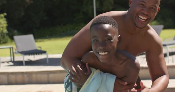 African American dad approaching, hugging teenage son holding towel, in trunks and posing at pool