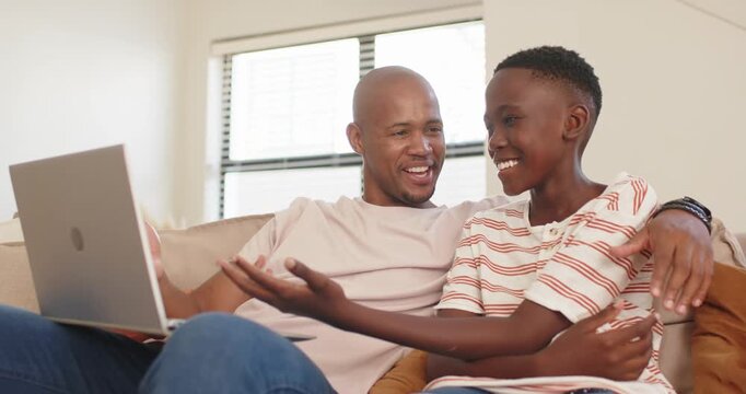 African father and youth son leaning closer, reacting to laptop content on couch at home, bonding