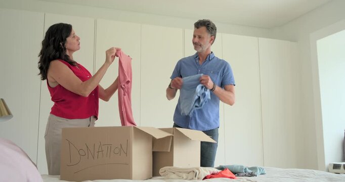 Couple sorting shirts on bed after closet cleanout, packing into box labeled DONATION for charity