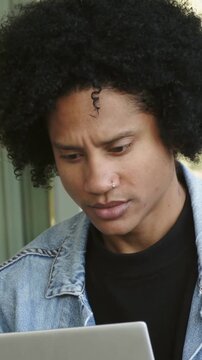 Vertical video: Camera pull causing man in denim leaning at door reading laptop adjusting bracelets