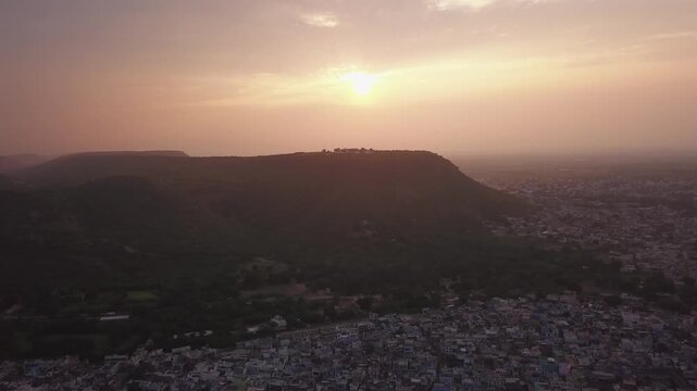 Aerial view at golden hour reveals Bundi's traditional blue houses and Taragarh Fort silhouette against warm sunrise sky. Atmospheric haze blankets the historic Rajasthan cityscape.