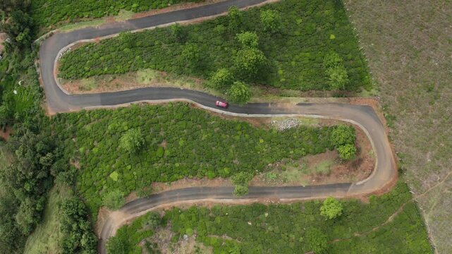Overhead drone view of a red car navigating a winding S-shaped asphalt road through hillside terrain near Ooty, Tamil Nadu. Green vegetation contrasts with cleared earth.