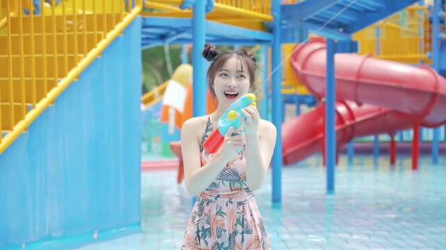 Young woman tourist in bikini swimsuit splashing water blaster spraying with water pistols or guns winking over blur park, toy gun festival holiday at a swimming pool. Concept of freedom enjoying life