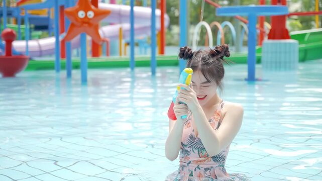 Young beautiful woman tourist in bikini swimsuit holding colorful squirt water gun, splashing water blaster spraying with water pistol or guns winking at water park swimming pool freedom enjoying life