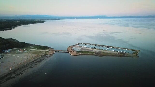 4K UHD drone flies backward over the calm, twilight waters of the Grado lagoon, revealing a small marina breakwater lined with moored boats, an access bridge, and the expansive lagoon horizon.