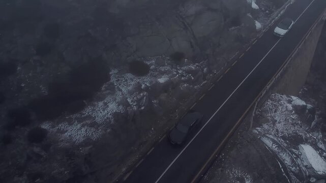 Gray car winds along N338 mountain road in Serra da Estrela, Portugal, cutting through rugged rocks and patches of snow beneath overcast skies in a dramatic alpine scene, top down drone tracking