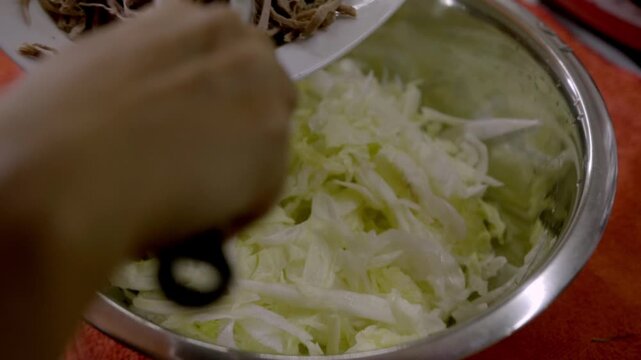 Close up of shredded meat being added into bowl with chopped cabbage and onions, mixing with utensils in kitchen