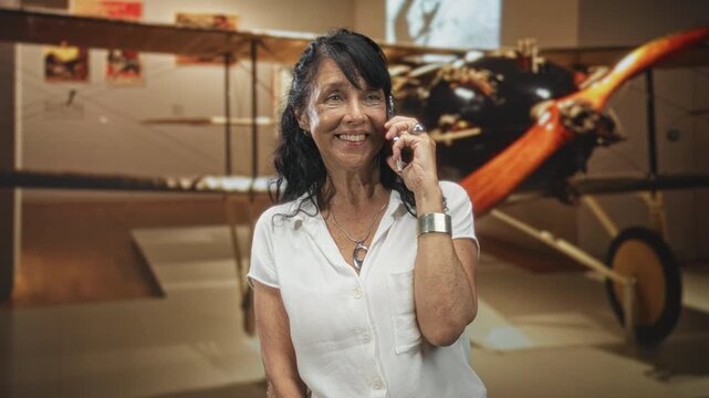 Woman talking on phone, smiling with phone to ear in a building beside a vintage biplane exhibit; joyful travel nostalgia.