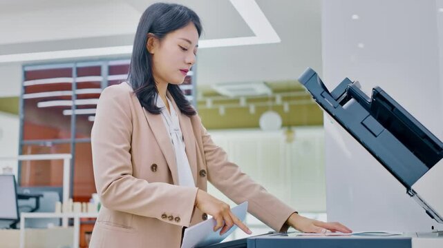Focused Asian businesswoman operating a multi-function printer, copying important documents in a bright, modern office environment for efficient workflow.