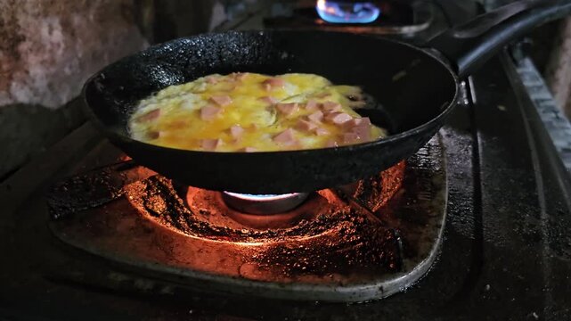 A basic meal of eggs and sliced sausage fries in an old, heavily worn pan over a bright blue gas flame. The extremely rusty and grease-stained stove highlights harsh, impoverished living conditions