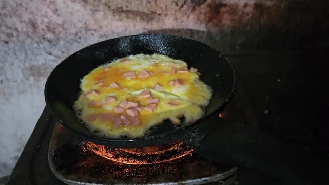 A simple, cheap meal of eggs and diced processed sausage is fried in a heavily worn, blackened pan over an old, rusty gas stove. The grimy and unhygienic conditions highlight the daily struggles