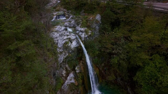 Pristine Ampola waterfall plunging into a turquoise natural pool surrounded by lush forest in the Ledro Valley, Italy. Aerial drone top-down view