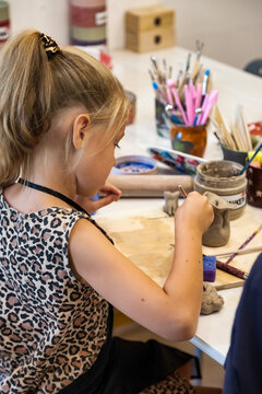 A young girl is diligently working with clay at a white table. She is shaping it with tools in a bright and creative workshop indoors. She is enjoying her time in an art class.