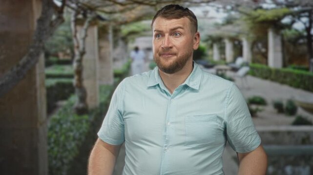 Man gesturing with hands in a building courtyard, seated in light blue short sleeved shirt with beard; casual doubt.