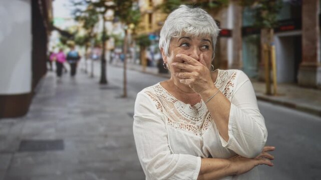 Woman plus size grey hair covering mouth with hand on street, arms crossed, eyes squinting in a suppressed laugh; amusement.