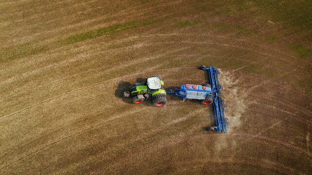 Aerial view of green tractor with blue plow working on brown agricultural field, creating furrows in soil during farming season
