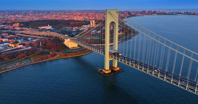 Numerous cars move on and from the Verrazano-Narrows Bridge in New York, USA. Distancing from the waterfront lit by the setting sun.