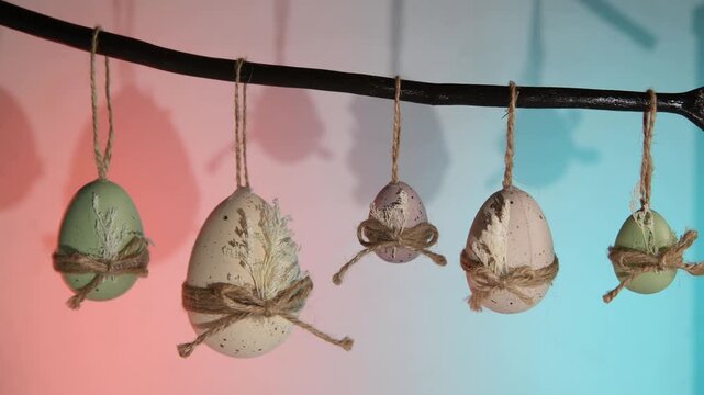 Pastel easter eggs in green, white, and purple hang from a natural branch, tied with twine bows and dried leaves against a vibrant background for spring celebration