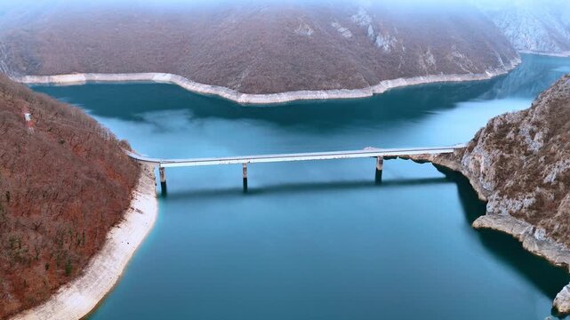Concrete bridge crossing Piva Lake between steep mountain hills. Aerial view of a long bridge over turquoise water connecting two slopes with autumn vegetation in Montenegro.