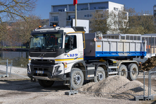 Nancy, France - April 2nd 2026 : View on a white Ampliroll truck Volvo FMX 460 with a removable grey and blue roll-off skip parked on a construction site with trees and buildings in the background.