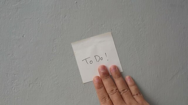Hand Sticking a 'To Do' Sticky Note on a Textured Grey Wall for Productivity and Task Management