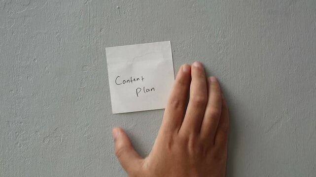 Hand sticking a sticky note with Content Plan written on it on a grey wall