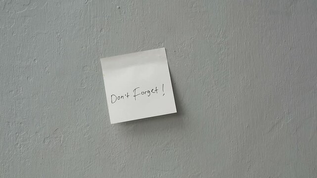 Hand placing a 'Don't Forget!' sticky note reminder on a grey textured wall