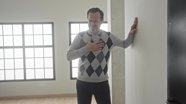 Middle aged man clutch chest with one hand, leaning on wall near window and pillar, grimacing and showing heartattack in building; pain emergency.