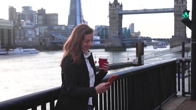 A young woman talking on her mobile phone and holding a coffee cup in front of the city skyline of London as a concept for business