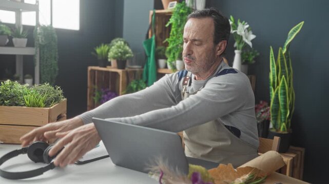 Man florist in apron with hands behind head stretching at laptop and bouquet on table, headphones and potted plants visible in studio; relief rest.
