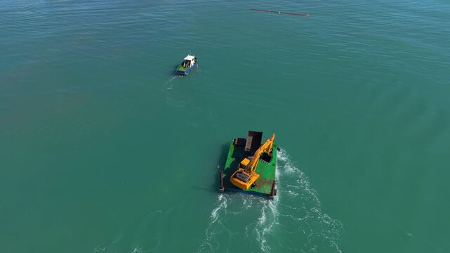Overhead view of a yellow excavator mounted on a green barge operating in turquoise ocean water, with a small patrol boat nearby during marine construction work