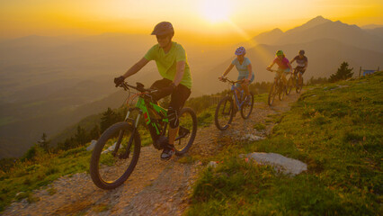 Obraz premium DRONE: Group of tourists riding electric bicycles up a scenic trail at sunset.