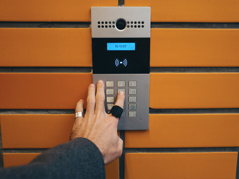 A man's hand enters a password on an intercom with a camera on an orange brick wall. Home security. Code lock.