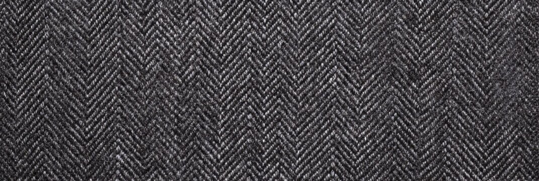 Detailed texture of a dark charcoal herringbone wool fabric with a traditional V-shaped weave. The textile surface shows intricate interlacing of black and grey threads in a banner format.