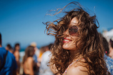 Naklejka premium Close-up of joyful woman with windblown hair at oceanfront party, people dancing blurred in background, bright blue sky, high detail, crisp natural lighting, advertising-ready composition, balanced