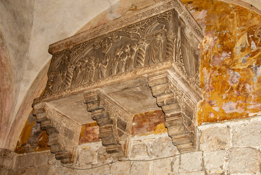 An intricately carved medieval stone corbel featuring detailed relief sculptures of robed figures, set beneath a weathered arch in ancient stone building. Croatia, Dubrovnik