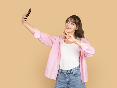 Smiling caucasian woman taking selfie with smartphone and making peace sign gesture, posing cheerfully concept, wearing pink shirt and denim, isolated on beige studio background with copy space.