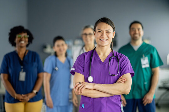 Portrait of a confident female nurse in purple scrubs with arms crossed, standing in front of a diverse medical team in a hospital setting, celebrating International Nurses Day