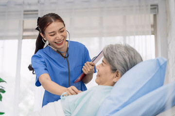 Fototapeta premium Smiling young Asian female nurse in blue scrubs with stethoscope providing compassionate care to elderly woman patient lying in hospital bed, representing International Nurses Day
