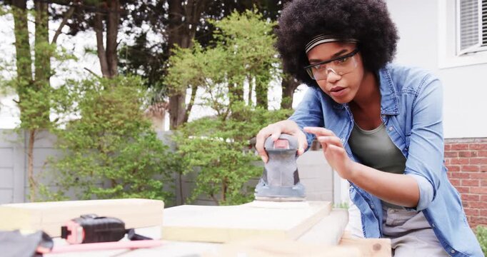 African American woman placing plank then sanding with orbital sander in backyard, smoothing wood